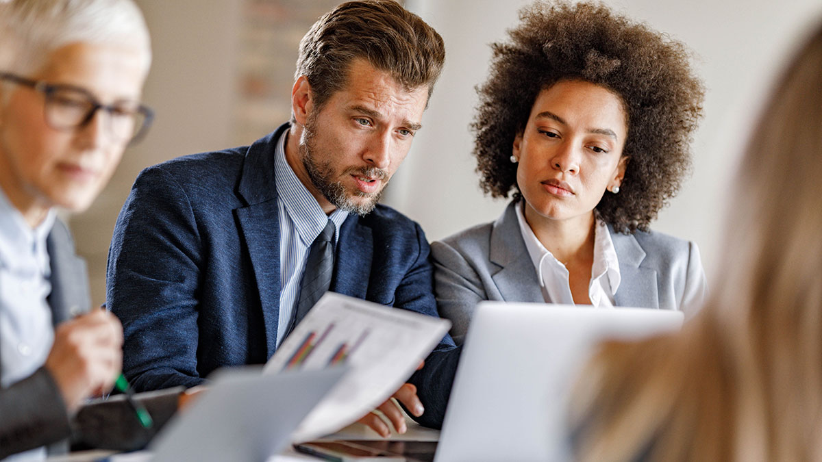 Three professionally dressed coworkers looking at a screen and analyzing paperwork.