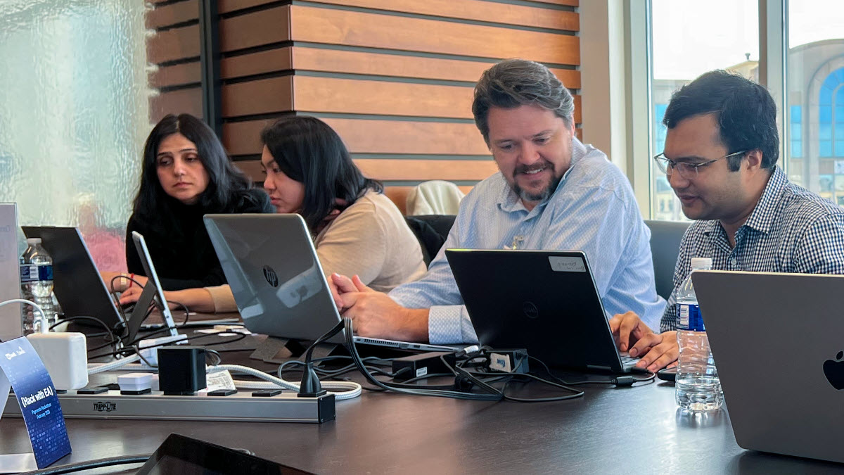 Four people sit by side-by-side at a conference table, reviewing information on their computers and talking to one another