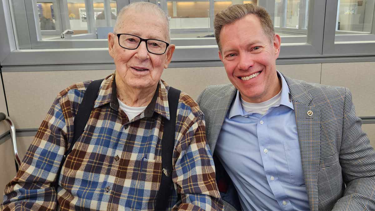 Older gentleman and younger man sitting on a couch smiling at the camera