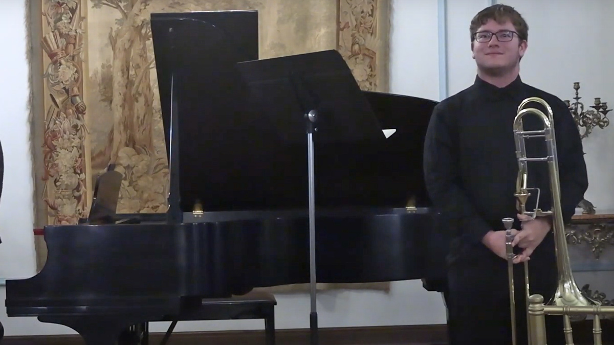 Young man holding a trombone and standing in front of a piano.