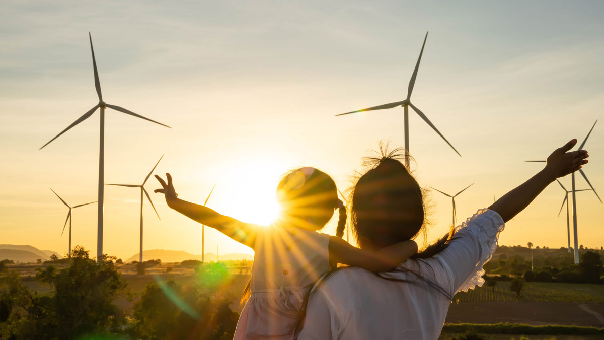 A woman and child from behind with their arms out, looking at the sun setting over a windmill farm.