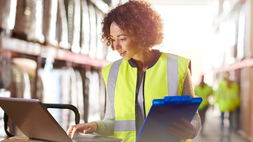 Person working on a laptop while holding a clipboard 