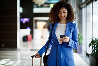 Businesswoman rolling a suitcase and using mobile phone