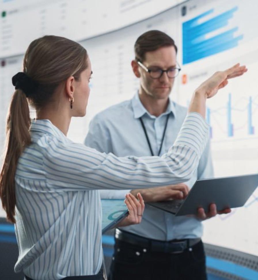 Two coworkers, a man and a woman, dressed professionally and standing in front of a large screen giving a presentation