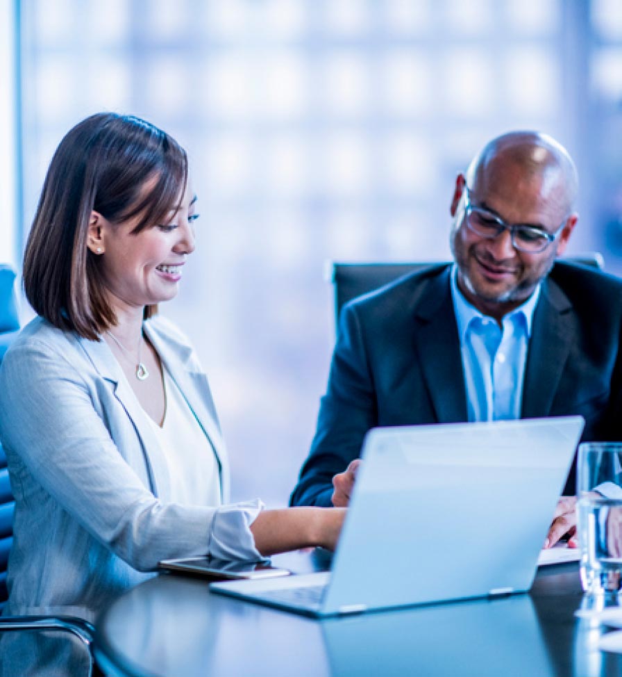 Man and woman, both wearing suits, seated at a large table and looking at a laptop together 