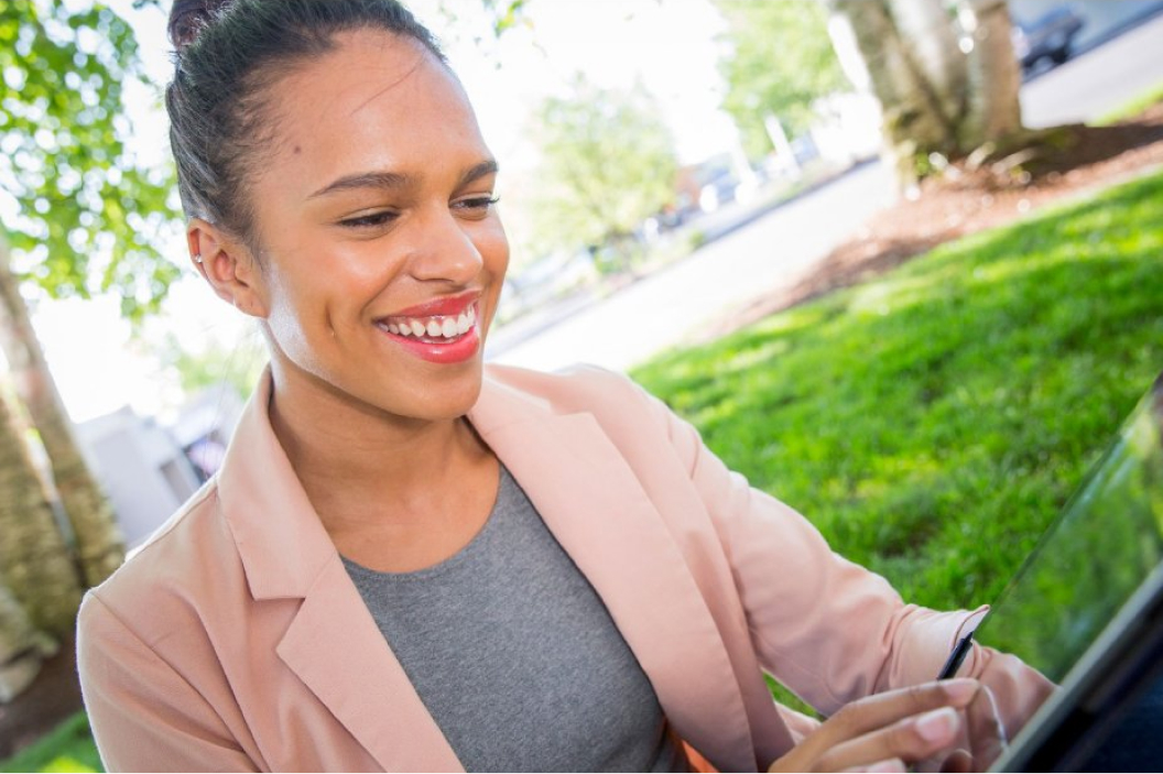 Woman wearing a light jacket in an outdoor setting. She is smiling while browsing the internet on her tablet device.