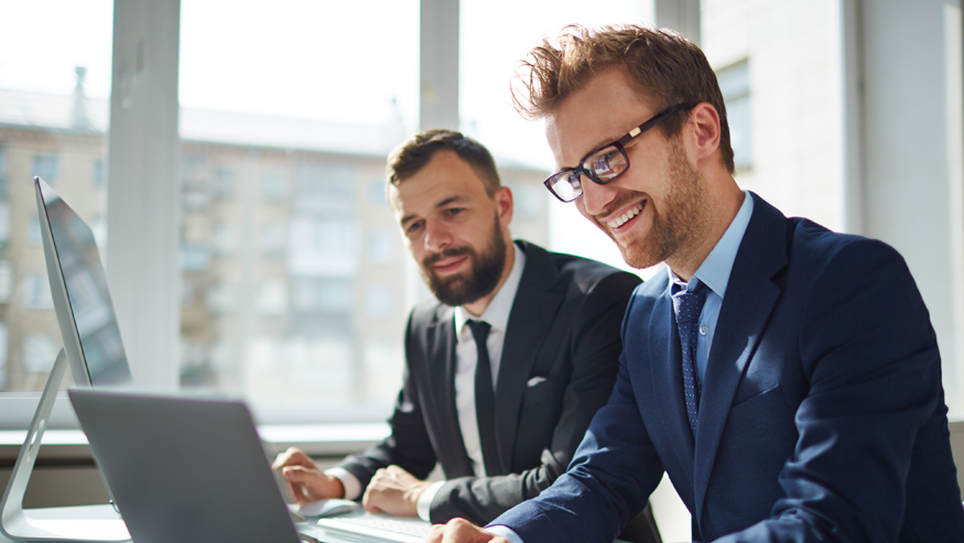 Two young professional men wearing suits and ties, sitting at a desk working on a computer.