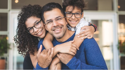 A Hispanic father stands in the middle, with his wife and child hugging him from behind.