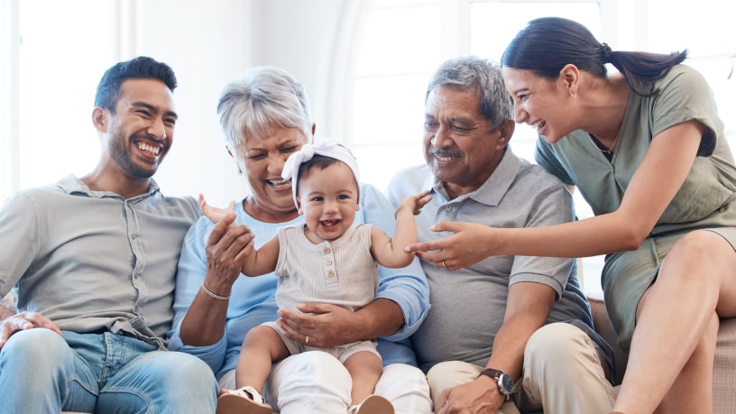 Three generations of a Hispanic family smile and laugh together, all the adults focused on the young toddler on her grandma’s lap.