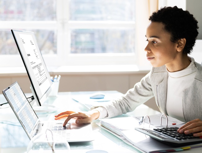 Professionally dressed woman at her desk with two computer monitors and a calculator.