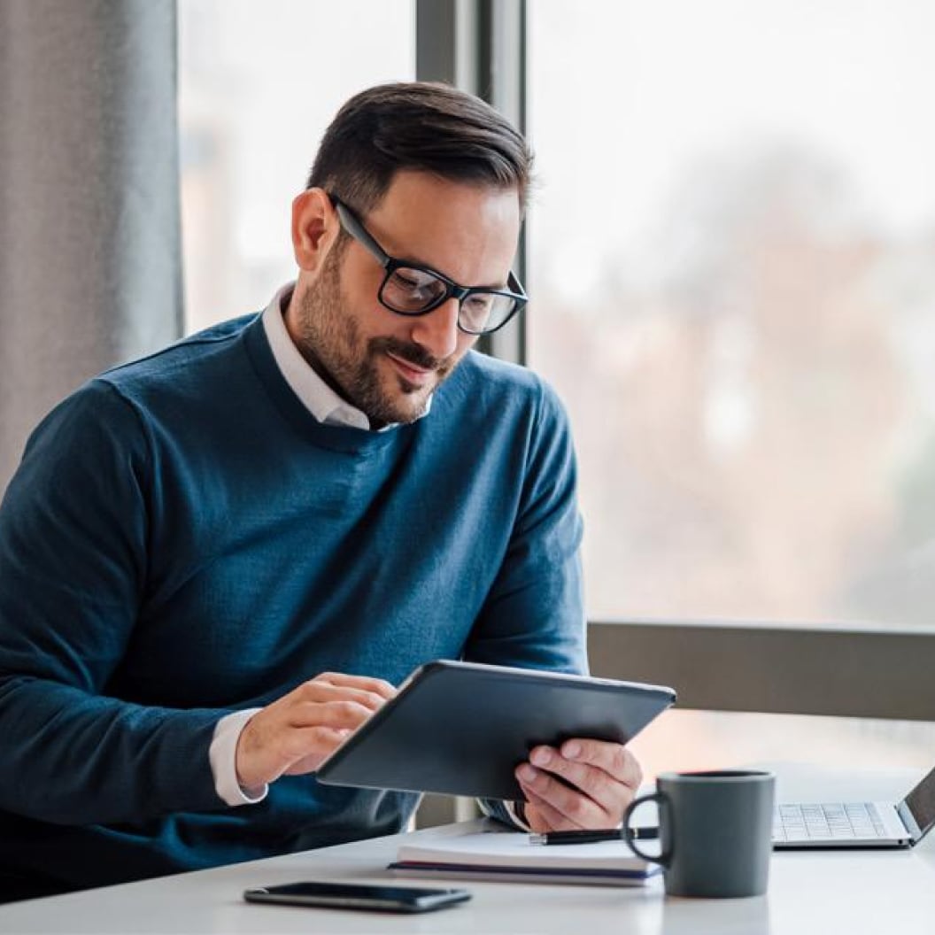 Man wearing glasses seated at a desk and looking at a tablet device and he also has a notebook and cup of coffee on the desk.