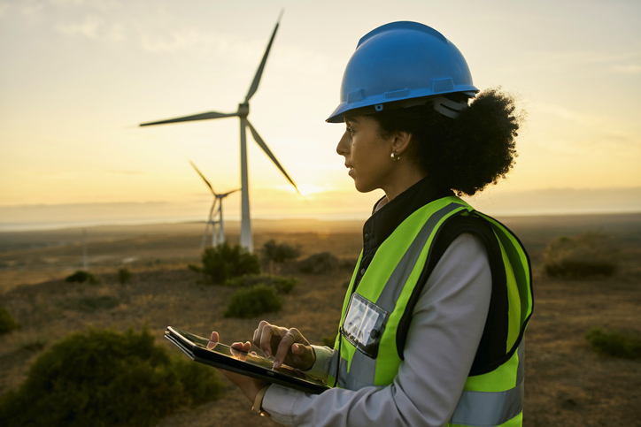 Woman wearing a hardhat standing near windmills on a wind farm. 