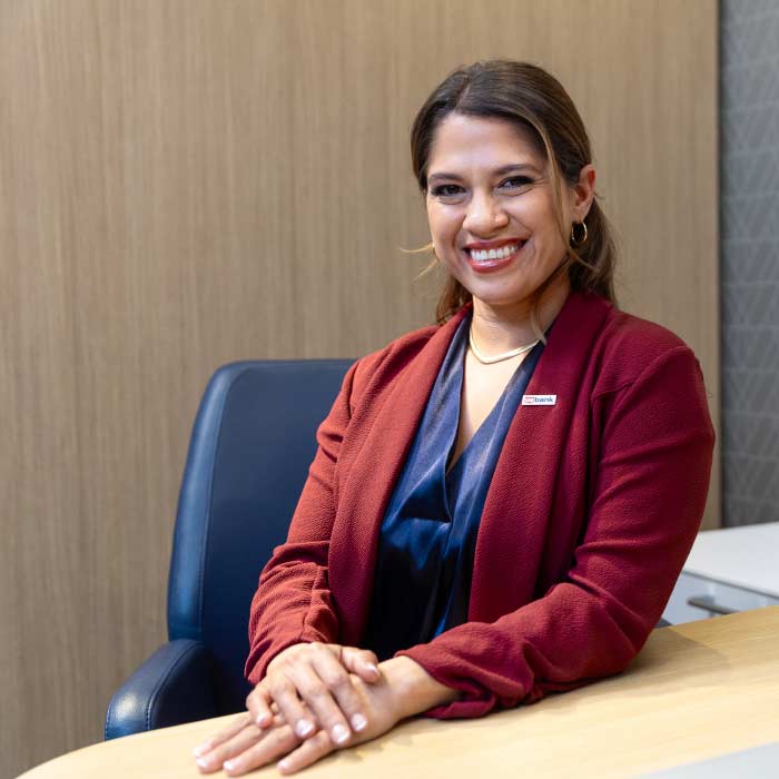 Mujer hispana joven sentada con una insignia del U.S. Bank en un escritorio, sonriendo para la cámara.