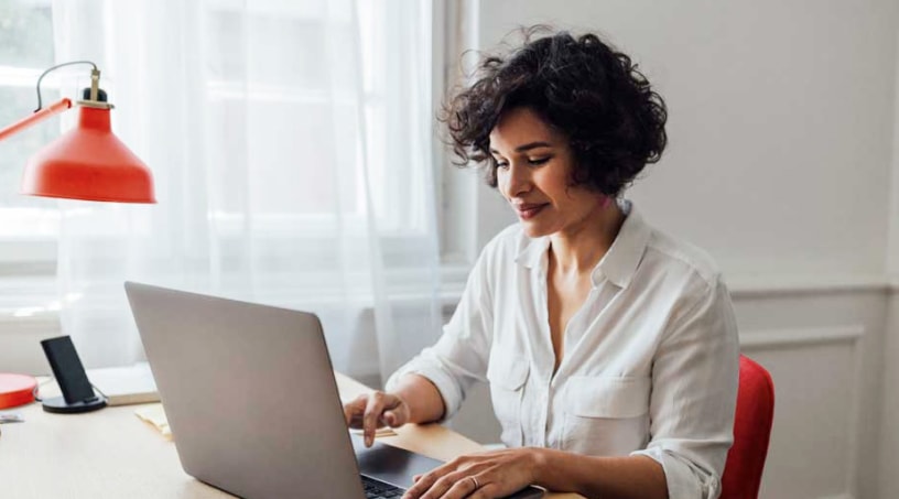 A young, brightly-dressed Black woman smiles at her laptop screen.